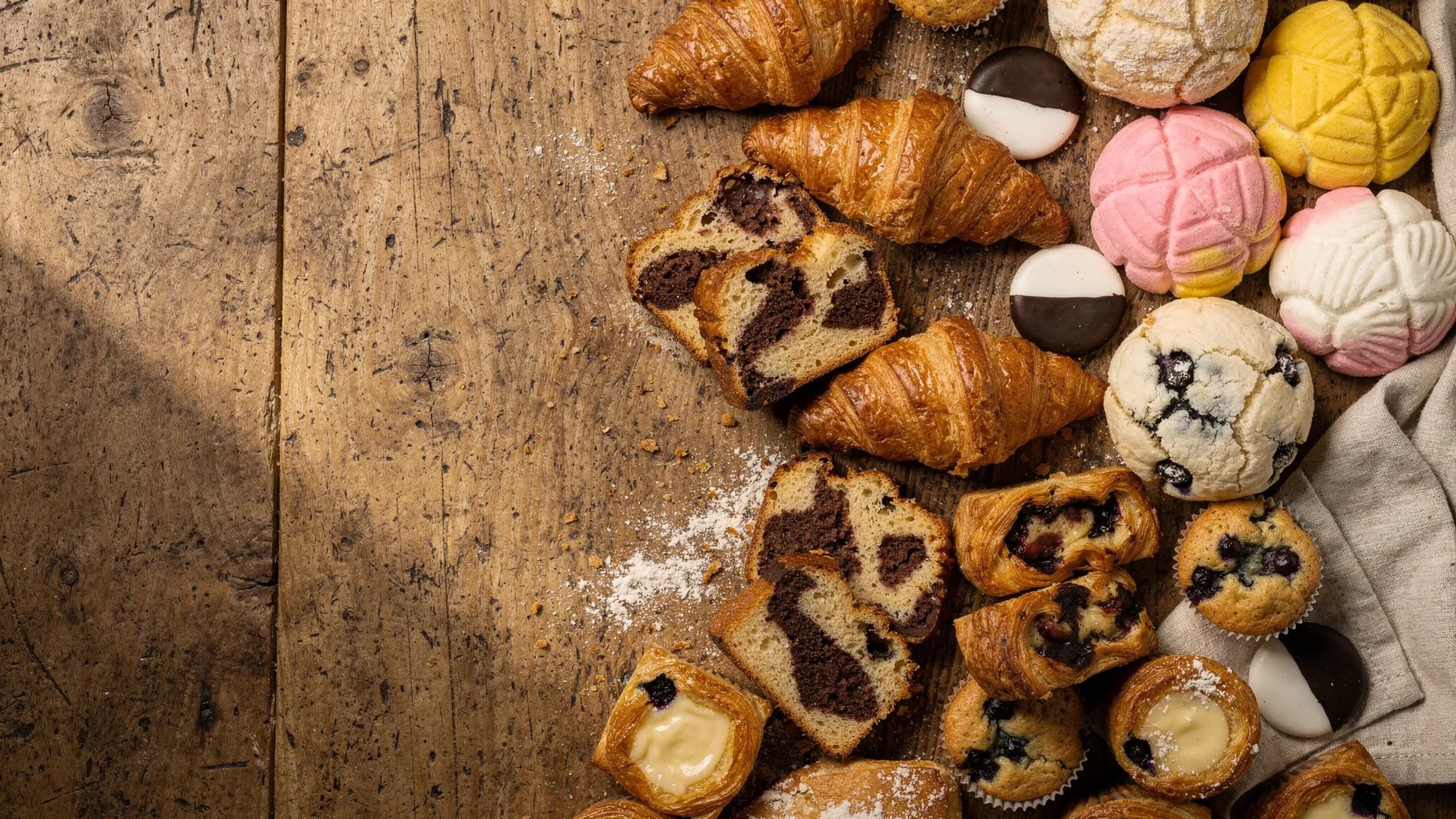 A spread of fresh baked goods on a long wooden table: croissants, black and white cookies, conchas, marble pound cake, muffins, and danishes